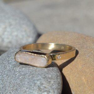 Oryginalny złoty pierścionek o surowym wykończeniu z nieszlifowanym kunzytem i małym, różowym diamentem w obrączce leży na naturalnym podłożu. Kamień główny ma ustawienie horyzontalne.