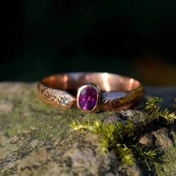 Prosty pierścionek z różowego złota o nierównej, nieregularnej powierzchni z owalnym, błyszczącym rubinem i małym diamentem w obrączce leży na mchu.