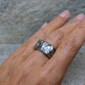 Duży rzeźbiony pierścień ze srebra leży na naturalnym kamieniu. Sygnet jest masywny, surowy, przyciemniany, ma załamania, rysy, szczeliny i wypukłe, jasne miejsca.