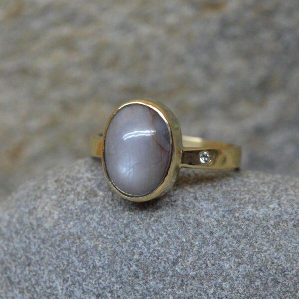 Prosty złoty pierścionek z szarym, surowym szafirem i małym szafirem w obrączce leży na popielatym podłożu. Kamień główny ma ustawienie pionowe i jest owalny.
