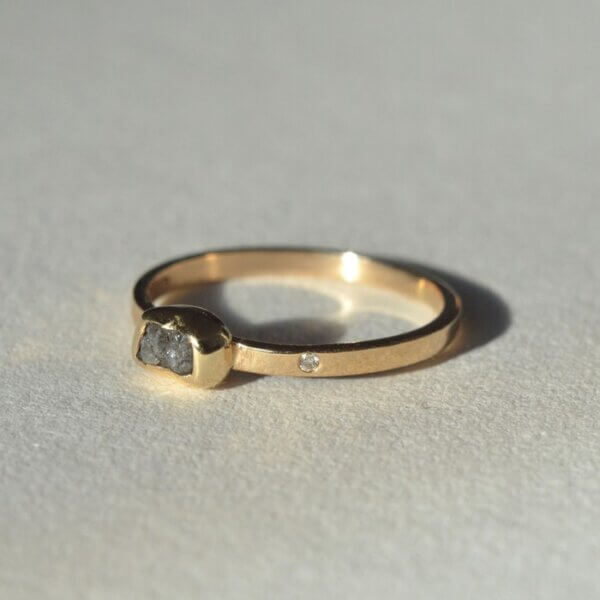 Elegancki złoty pierścionek o surowym wykończeniu z popielatym surowym diamentem i małym różowym brylantem w obrączce leży na szarym kamieniu