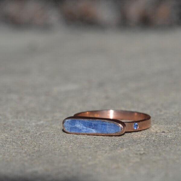 Prosty złoty pierścionek o surowym wykończeniu z niebieskim kamieniem i małym szafirem w obrączce leży na popielatym podłożu. Kamień główny ma ustawienie horyzontalne